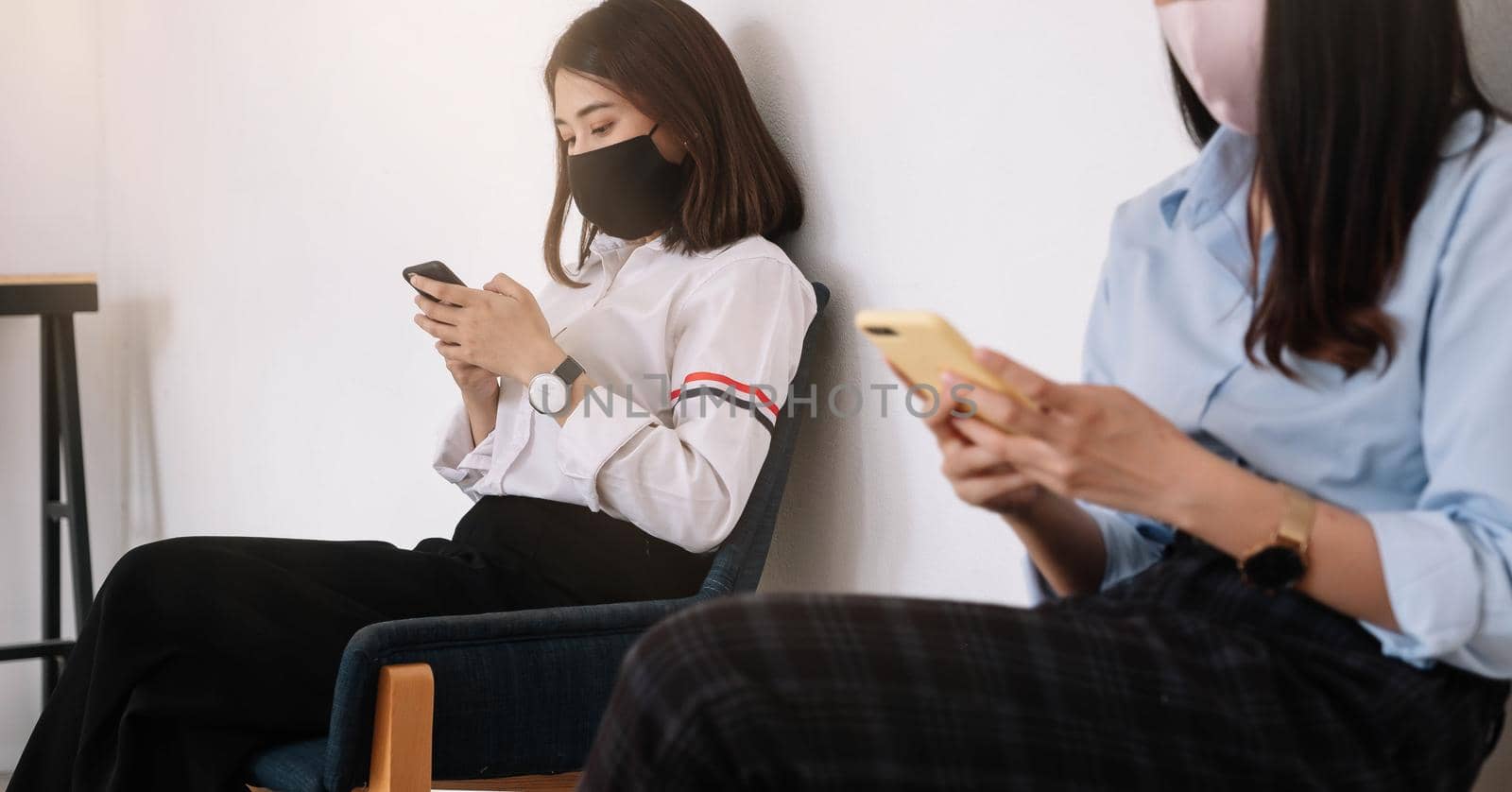 Two Asian young woman chatting in smartphone and wearing mask sitting distance protect from COVID-19 viruses for social distancing for infection risk. by nateemee