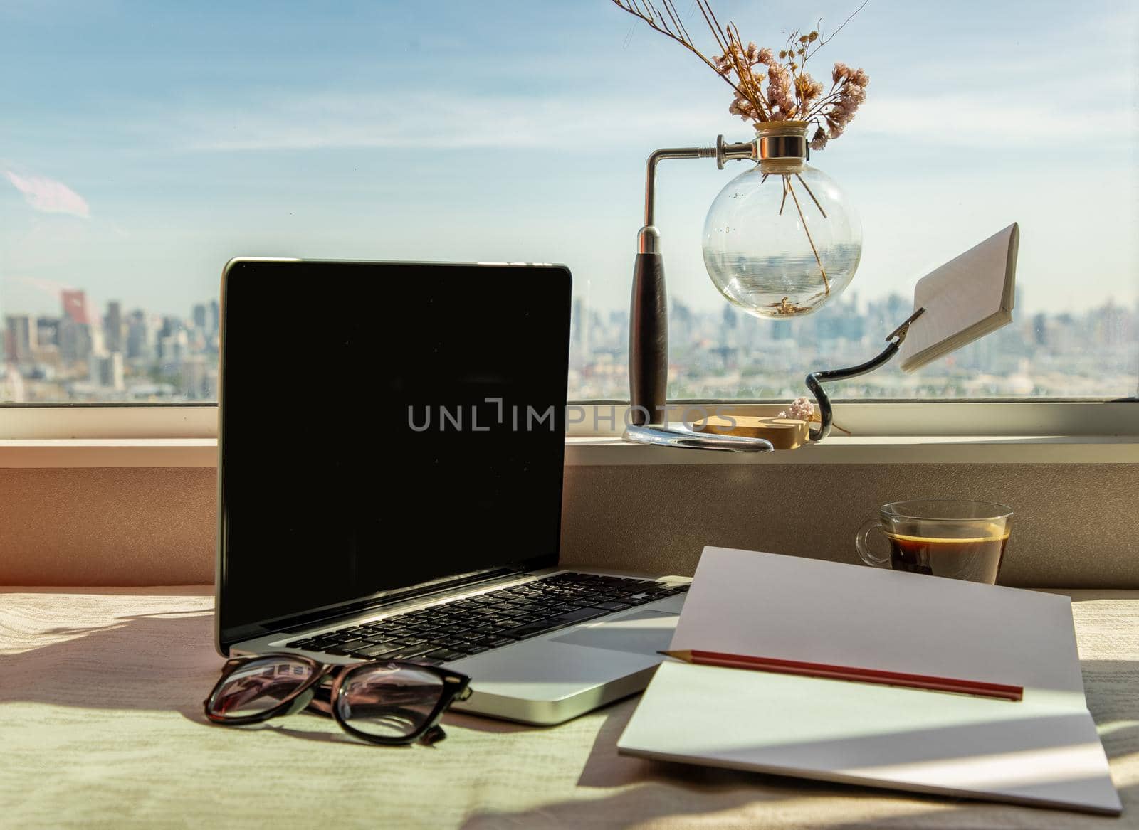 Working space with laptop, notebook, pencil, sunglasses, coffee cup, dried flower vase by the window with city views. Remote working concept or Business workspace at home or office.
