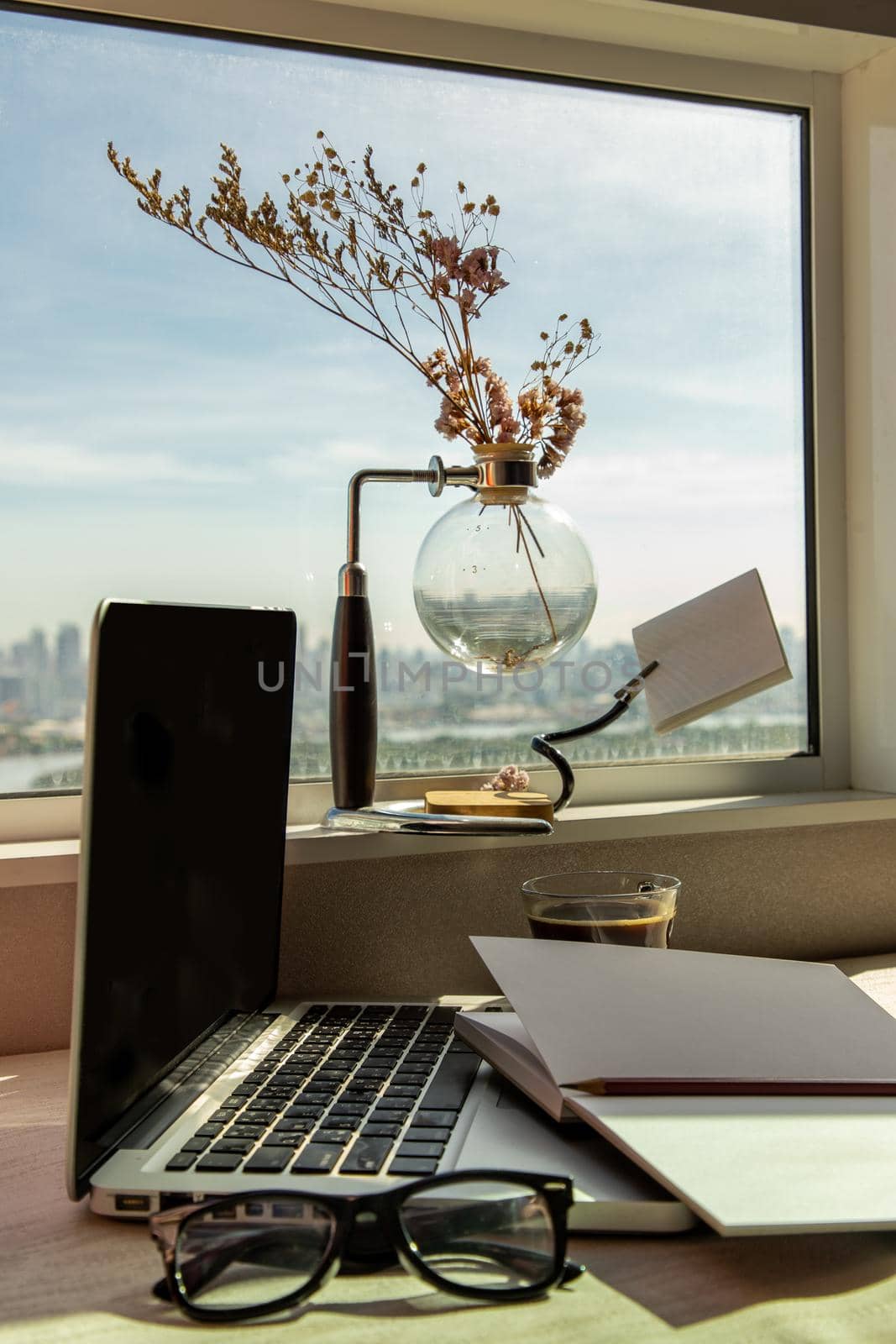 Working space with laptop, notebook, pencil, sunglasses, coffee cup, dried flower vase by the window with city views. Remote working concept or Business workspace at home or office.