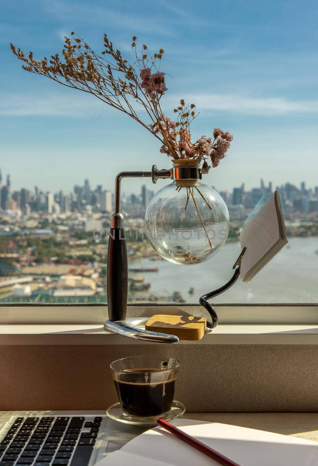 Working space with laptop, notebook, pencil, sunglasses, coffee cup, dried flower vase by the window with city views. Remote working concept or Business workspace at home or office.