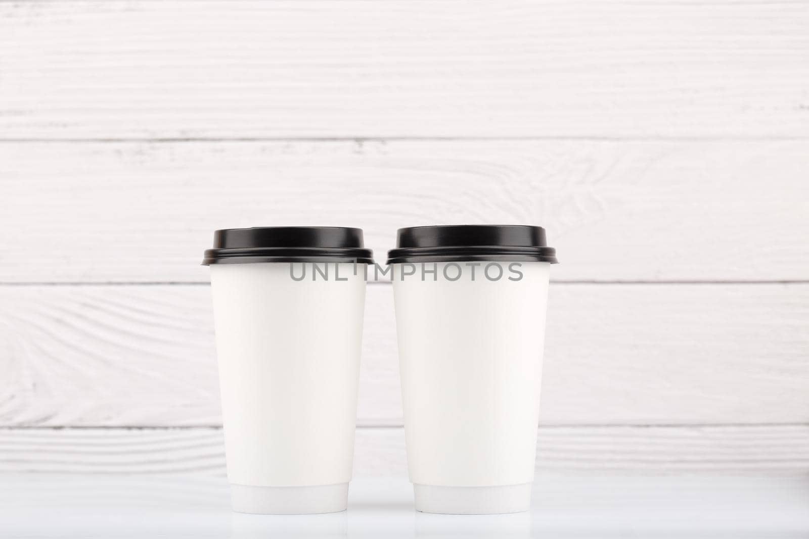 Two white tall disposable coffee cups on white table against white wooden background. High quality photo