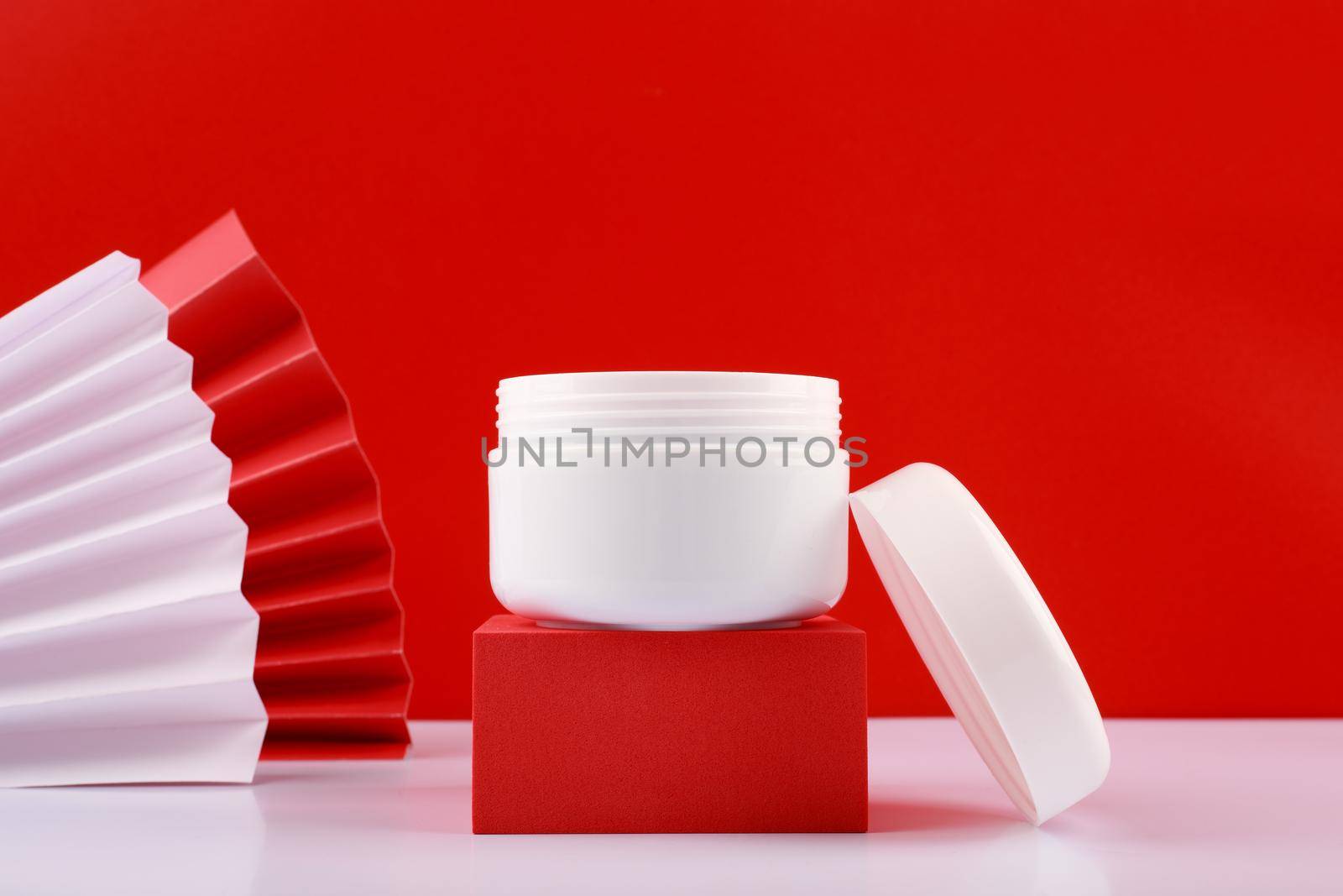 Selective focus, close up of white opened creme jar on red podium against red background decorated with red and white wavers. Concept of skin care and beauty