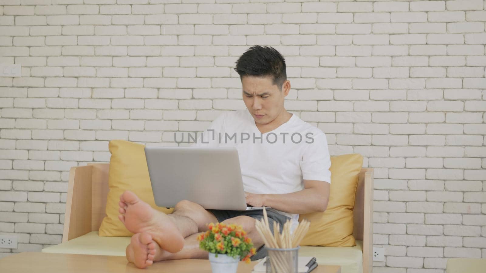 Happy Asian handsome man using and working with a laptop with online internet from home. Young man smiling reads the screen computer while relaxing on the sofa.