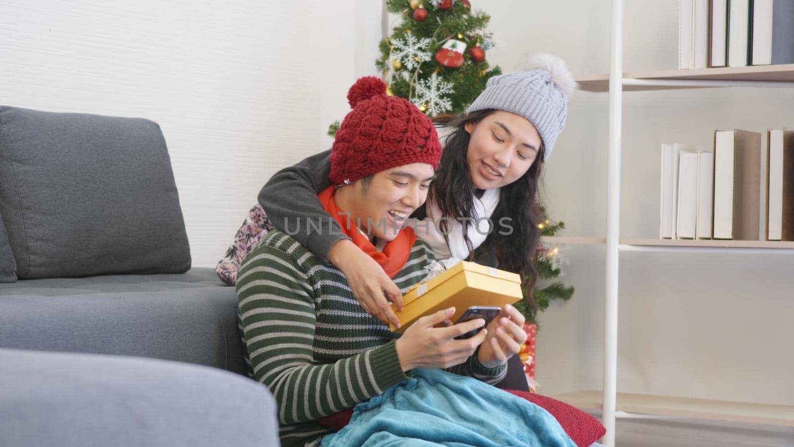 Happy Asian beautiful young family couple husband and wife in home living room, woman hide surprise giving gift box her boyfriend present during play smartphone, winter holidays and valentine concept