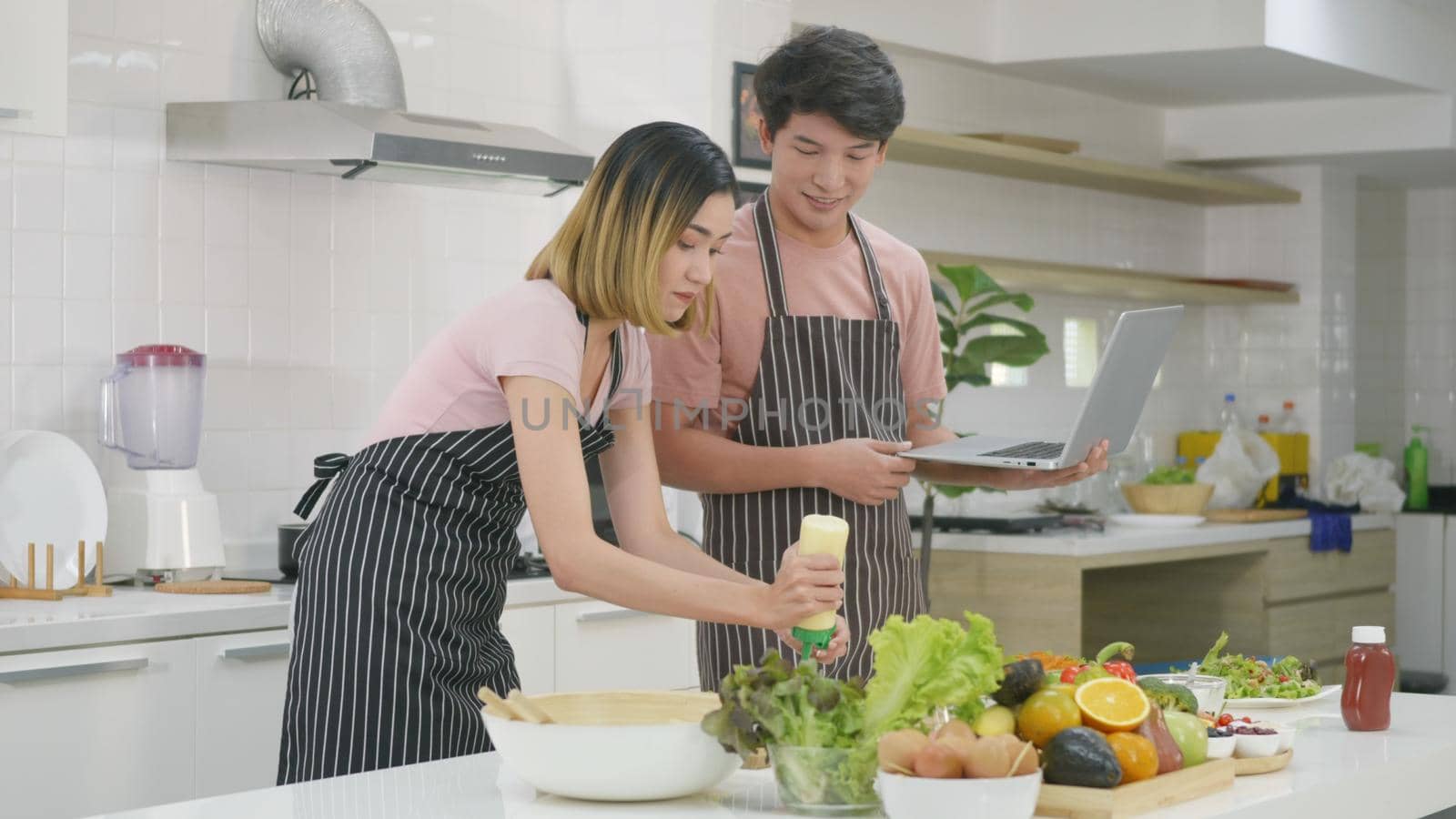 Happy Asian beautiful young family couple husband and wife cooking food vegetable salad in kitchen together at home. The man and woman according online cooking class to recipe on laptop computer