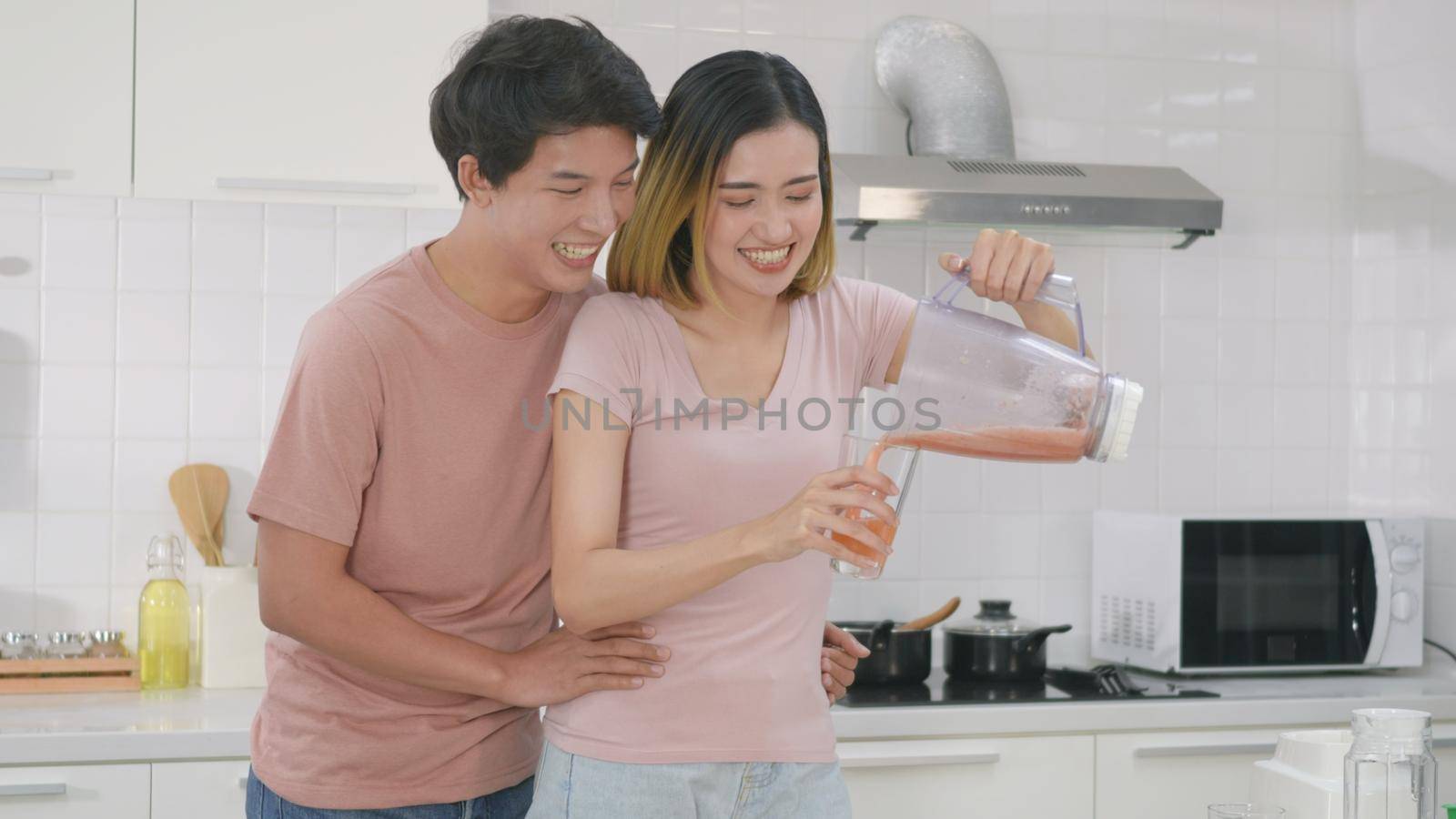 Happy Asian young family couple husband and wife enjoying drinking fresh apple smoothie juice in kitchen at home. woman poured juice from blender for man to drink. Healthy lifestyle.