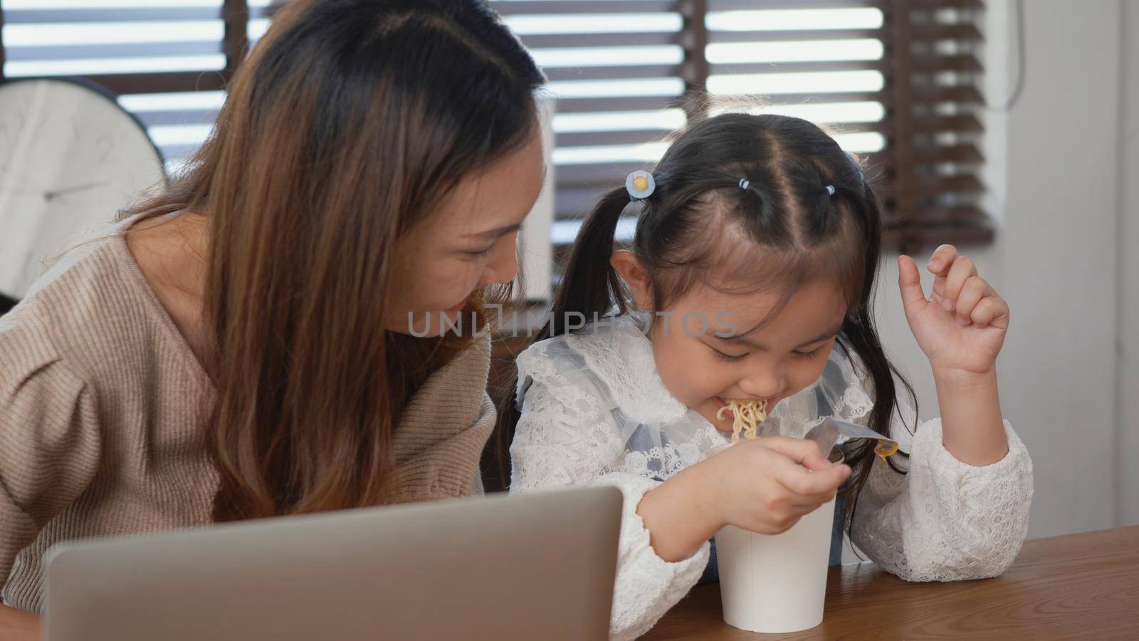 Asian family young mother and her little daughter smiling using laptop computer together for learn online and eating instant noodles at home, Child eats noodles while doing homework
