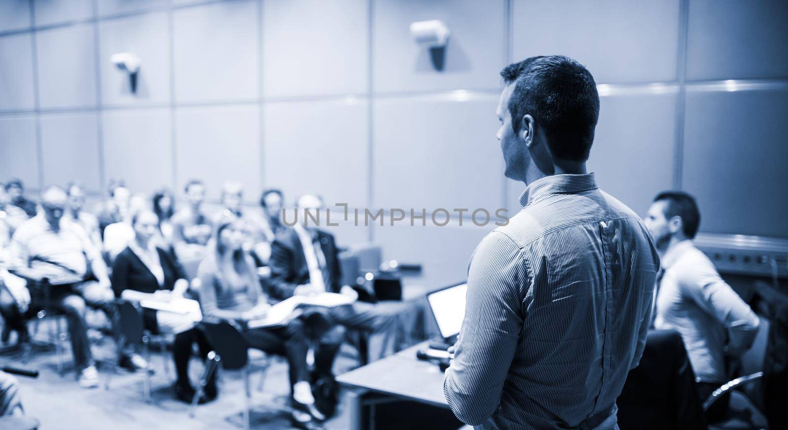 Speaker giving a talk on corporate business conference. Unrecognizable people in audience at conference hall. Business and Entrepreneurship event. Blue toned black and white.