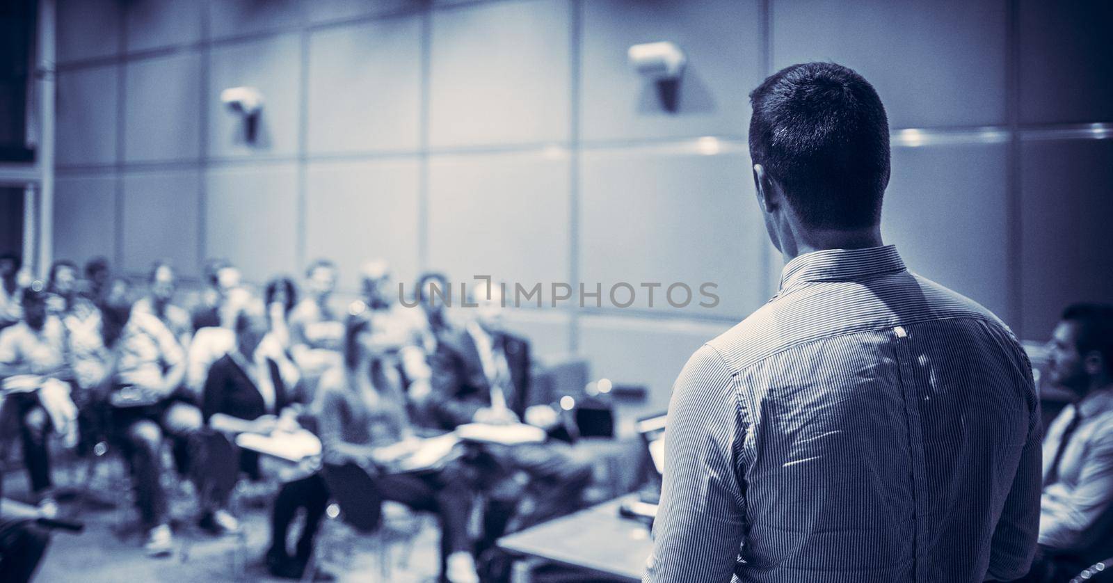 Speaker giving a talk on corporate business conference. Unrecognizable people in audience at conference hall. Business and Entrepreneurship event. Blue toned black and white.