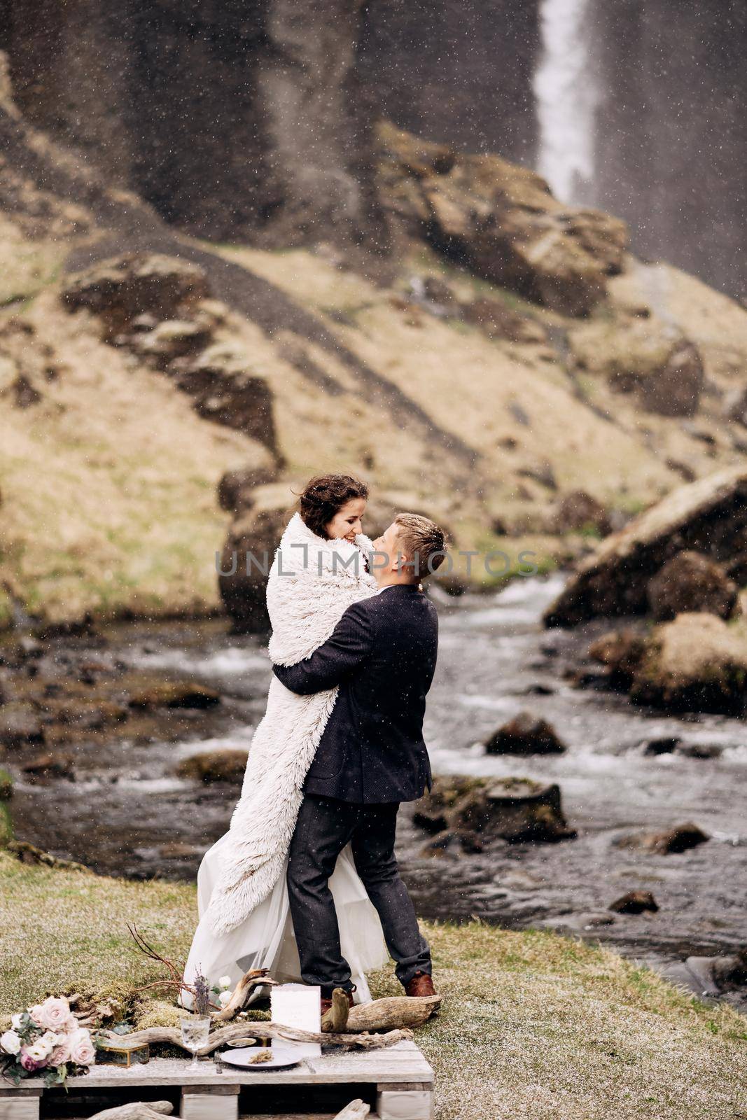 Wedding couple on the banks of a mountain river. The groom wears the bride in a woollen plaid in his arms.