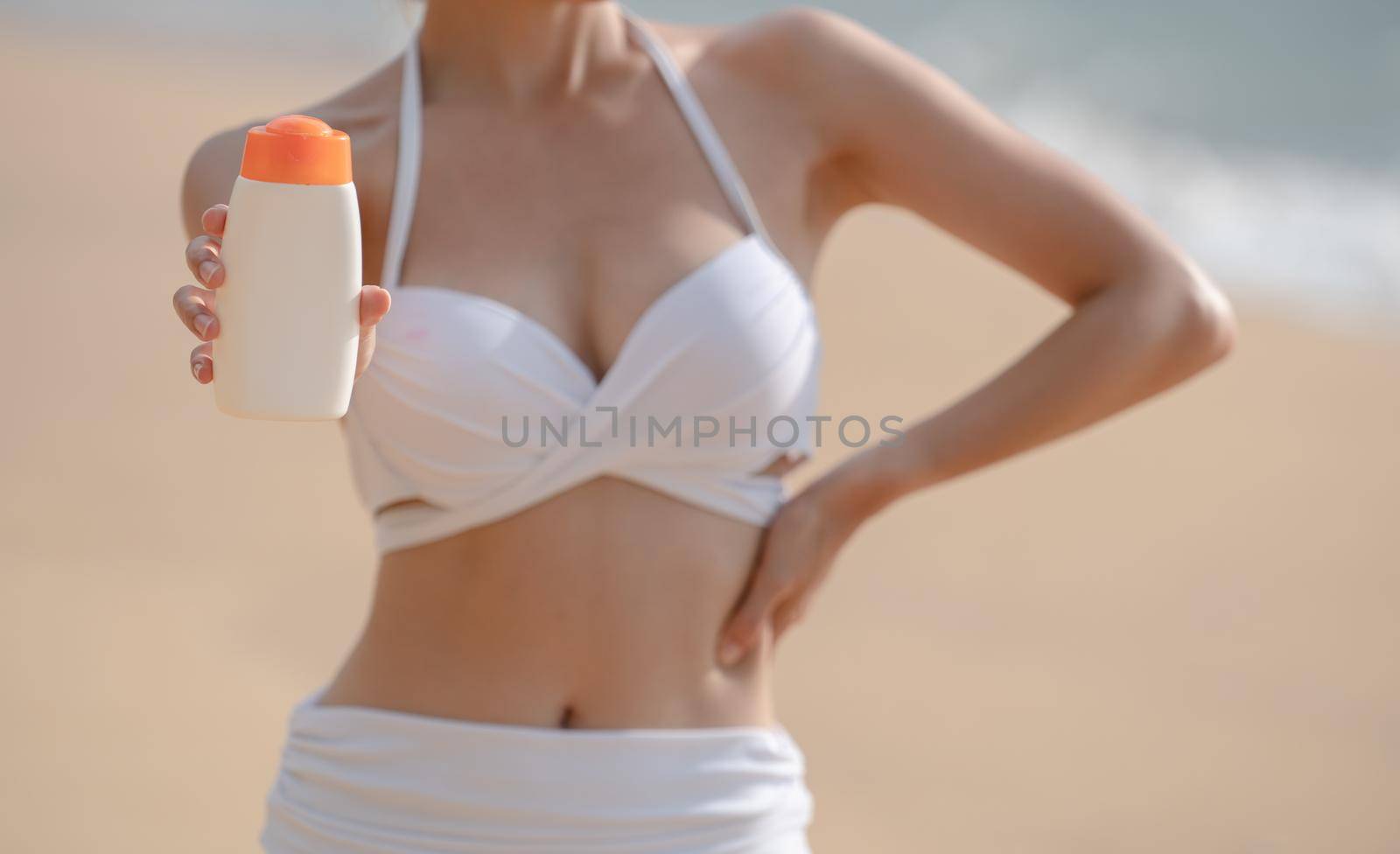 Woman in white bikini holding sunscreen bottle in hand on the beach. by sirawit99