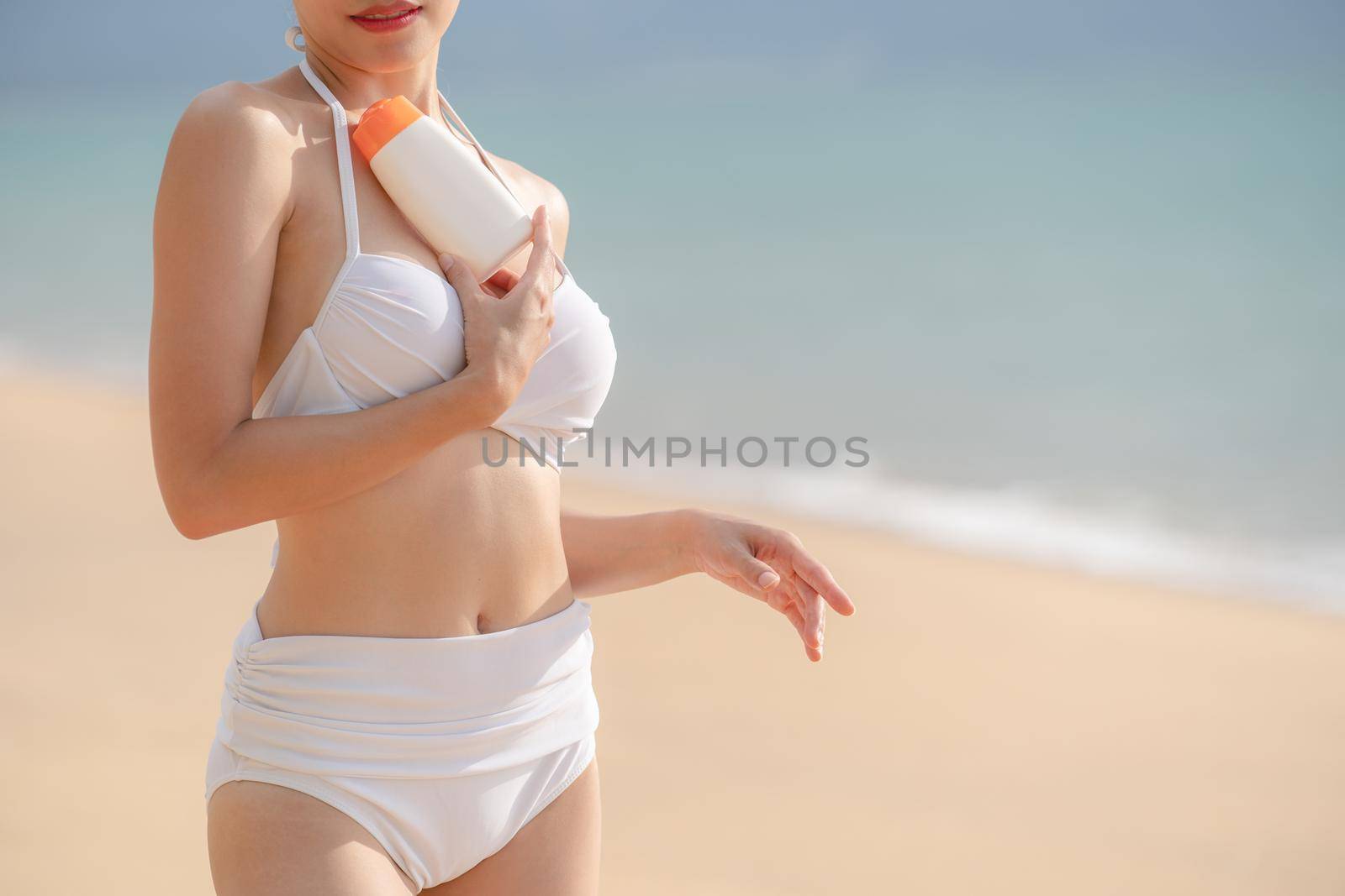 Woman in white bikini holding sunscreen bottle in hand on the beach. by sirawit99