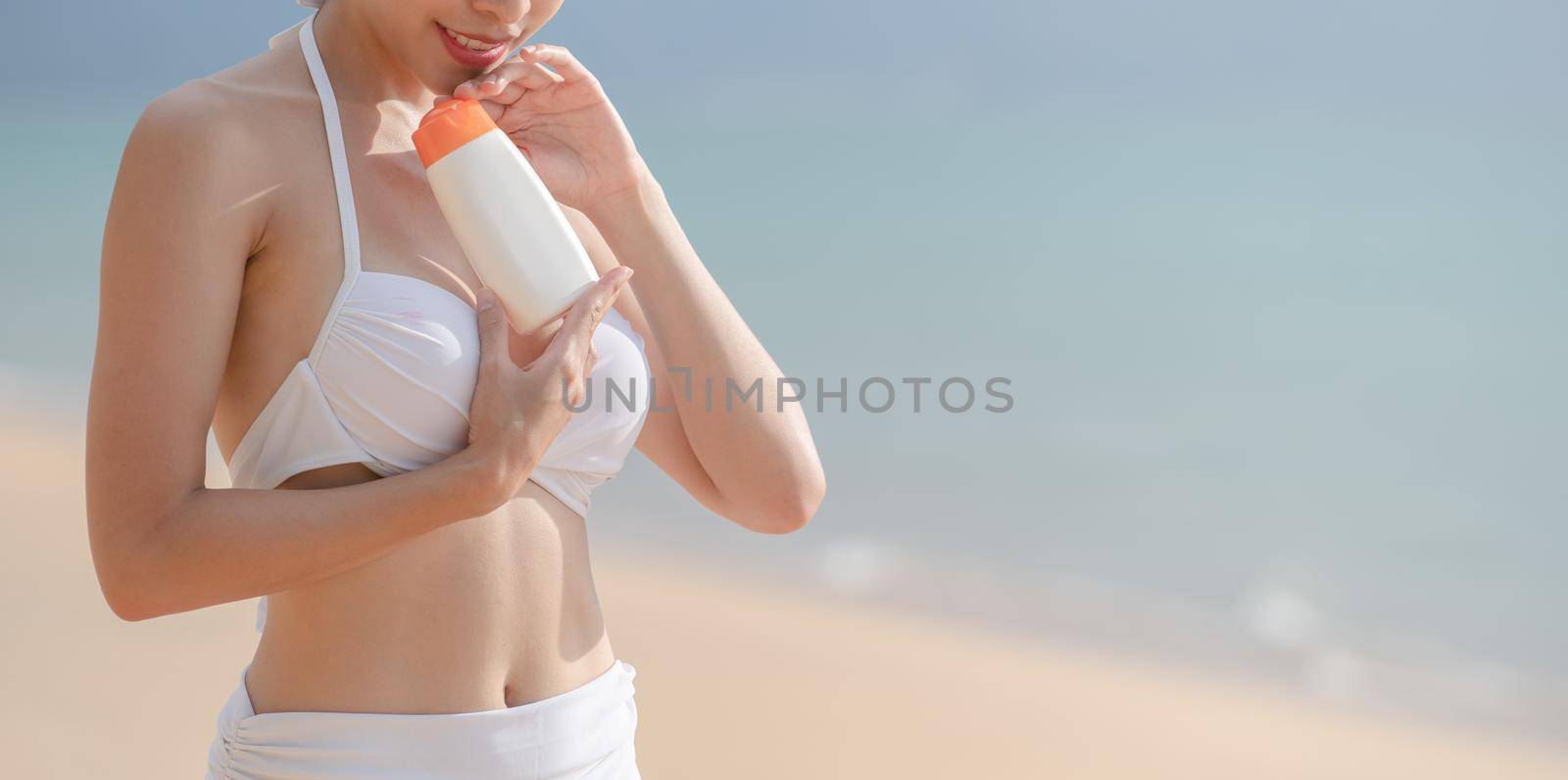 Woman in white bikini holding sunscreen bottle in hand on the beach.