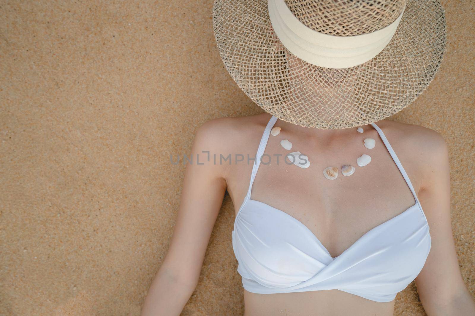 Woman in white bikini lying on sand beach making necklace from sea shell, straw hat covering her face.