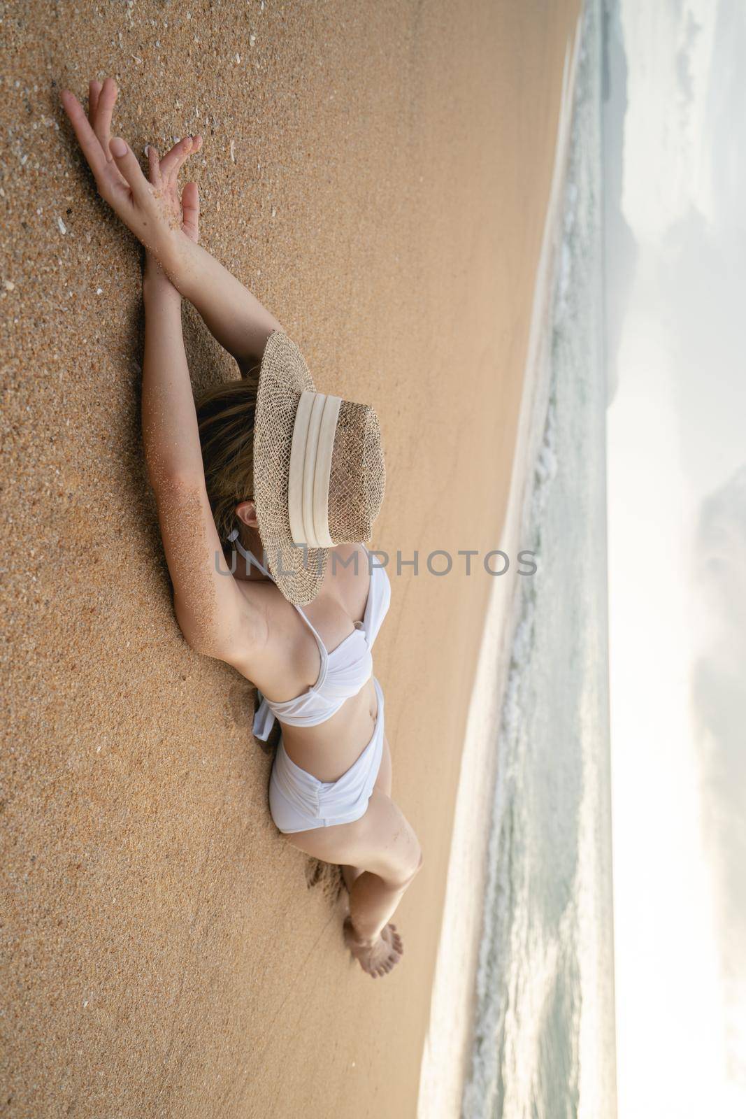 Woman in bikini lying down on sand beach with straw hat cover her face, relaxing sunbathing.