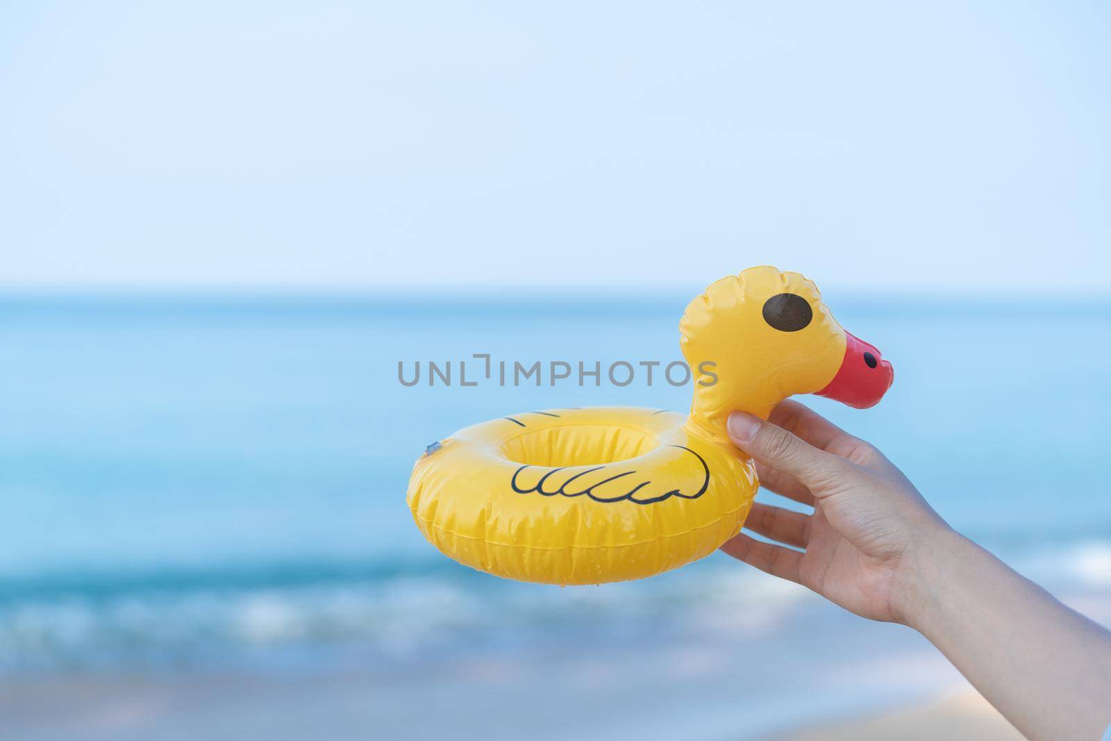 Hand holding a yellow duck on the beach with a sea in the background.