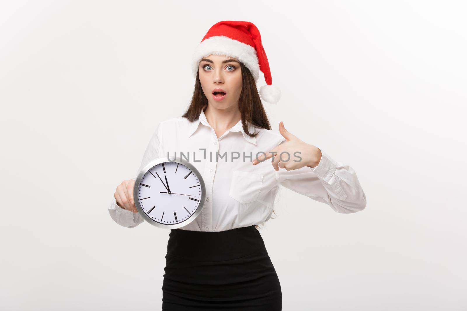 Time management concept - Young business woman with santa hat holding and pointing a clock isolated over white background. by Benzoix