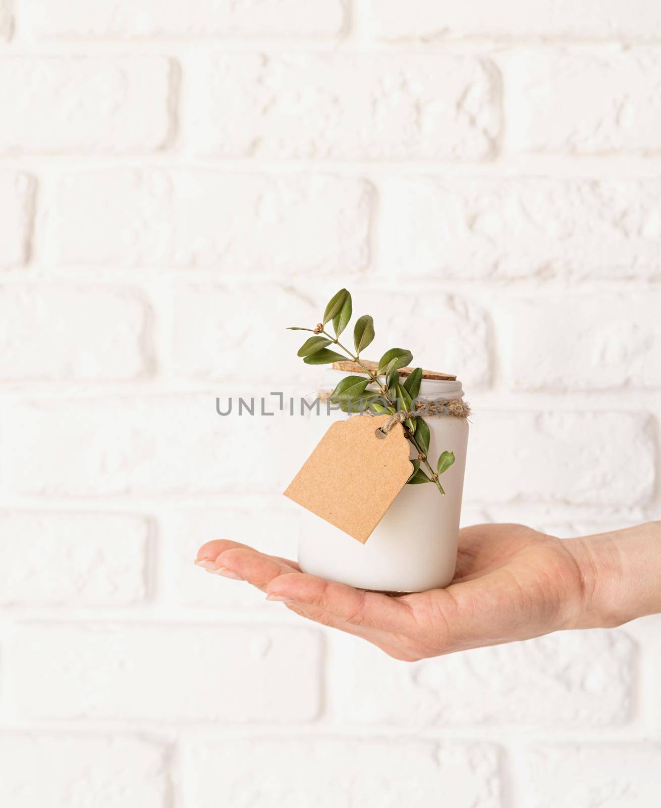 Home decoration and interior. Female hand holding beautiful white candle with fresh leaves on white brick wall background