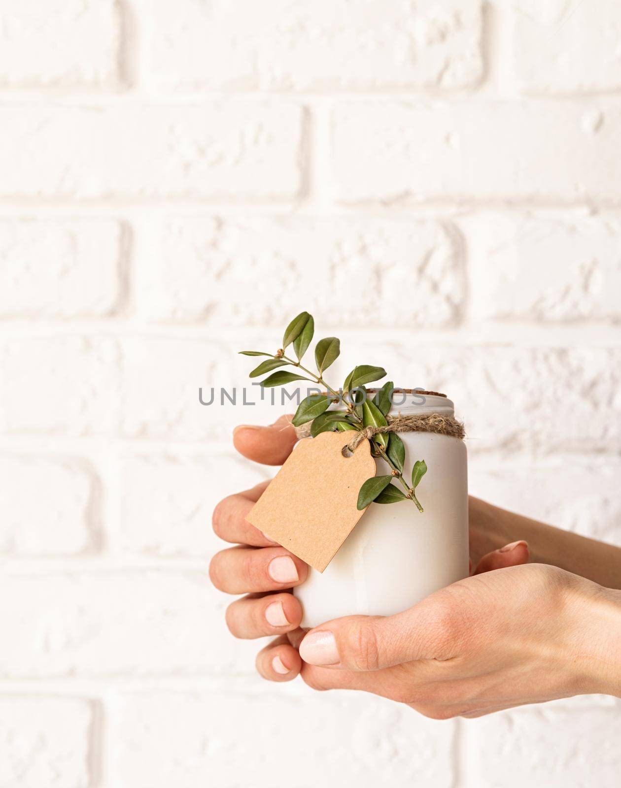 Female hand holding beautiful white candle with fresh leaves on white brick wall background by Desperada