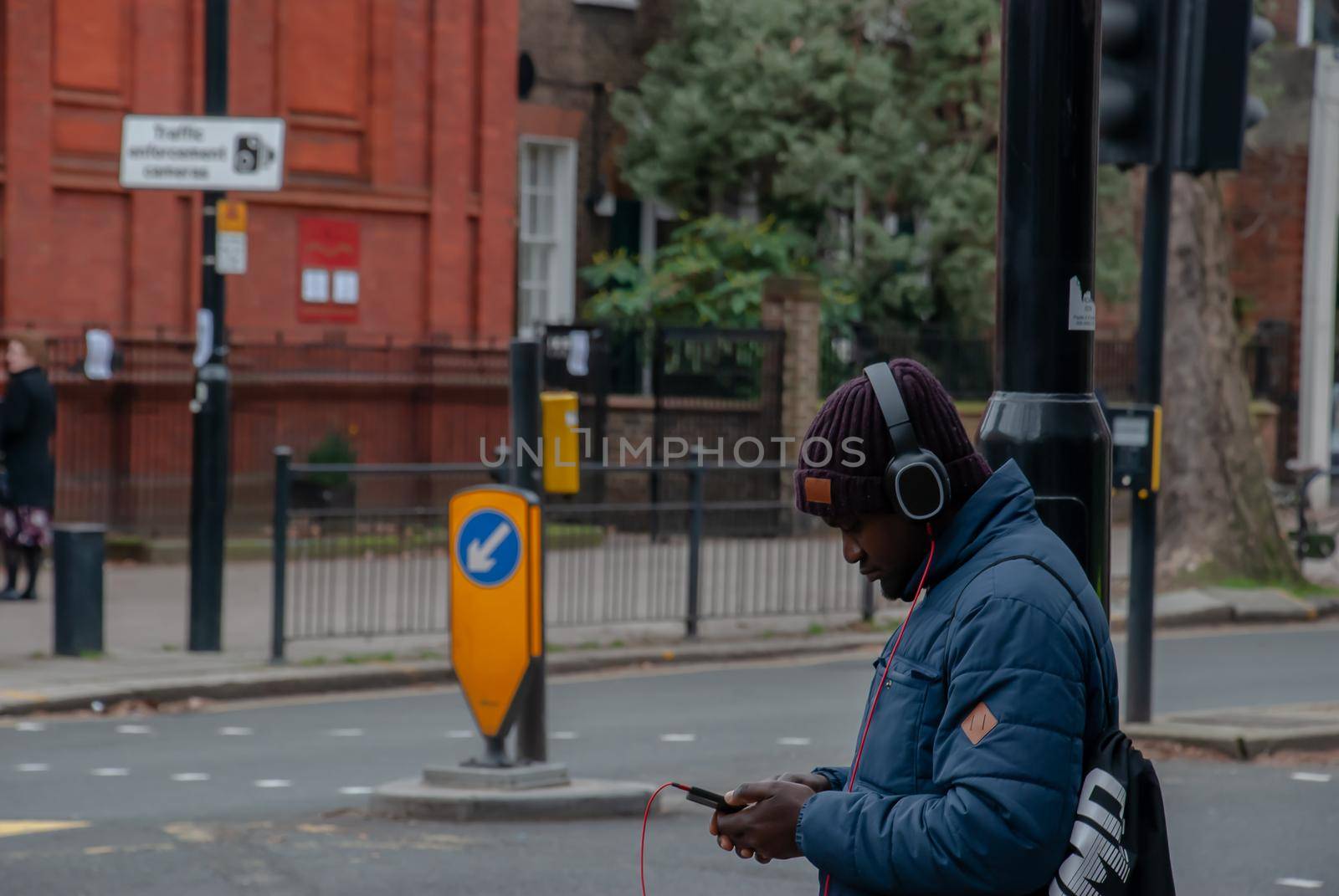 London, United Kingdom - APR 05, 2019 : A young black english man wearing black headphone and listening to music on his smart phone while waiting for the green light to cross the street of London.