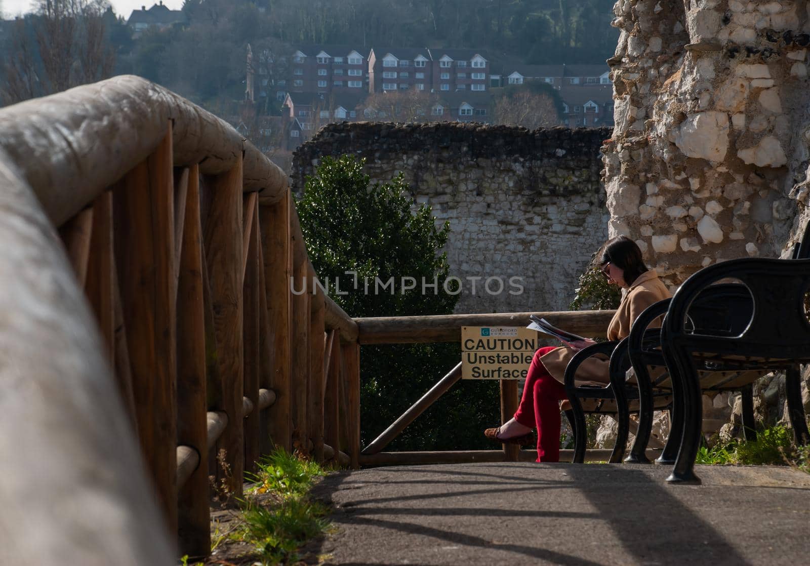 Guildford, United Kingdom - May 05, 2019 : A young woman sitting reading book on wooden bench in Guildford Castle and Grounds. No focus, specifically.