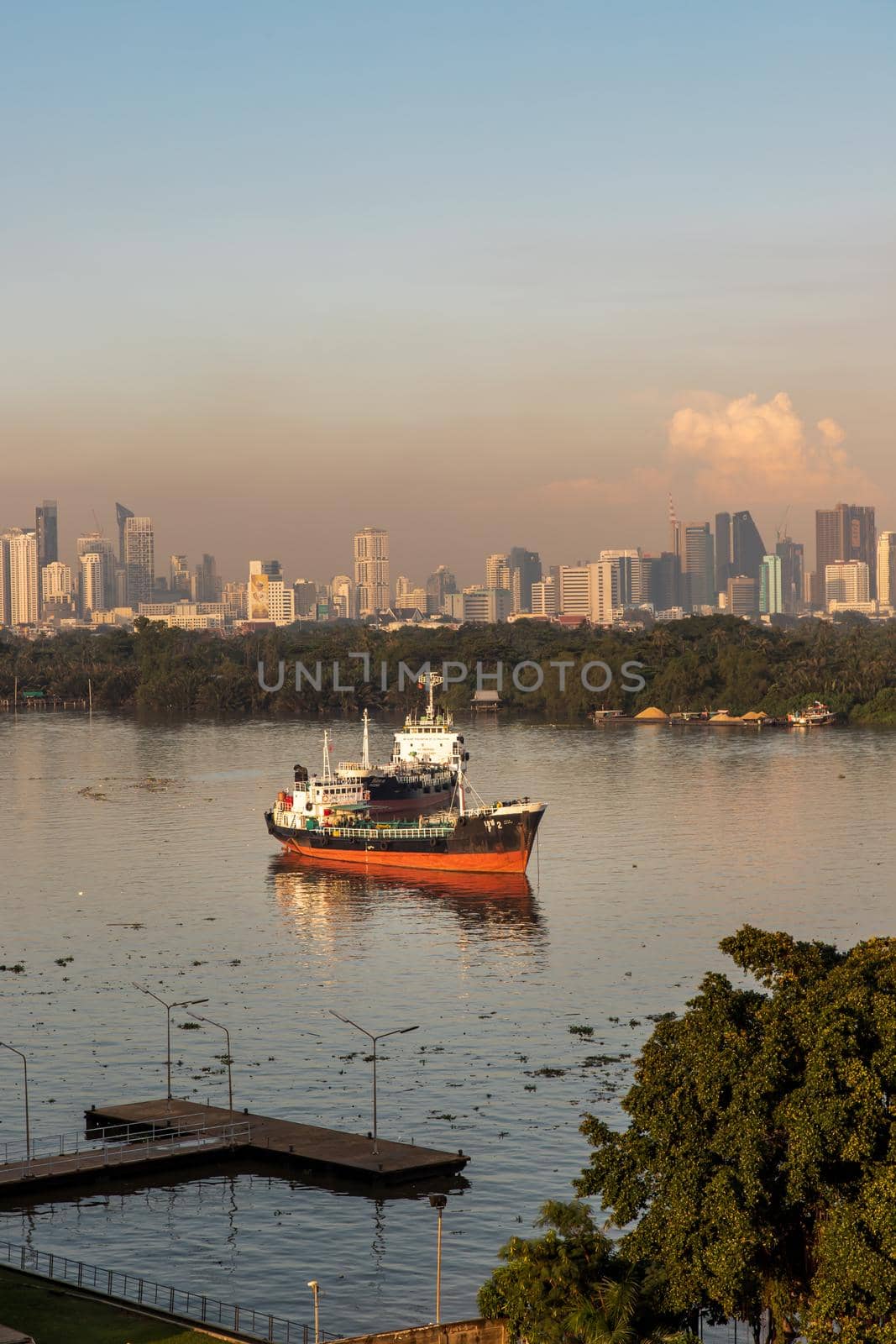 Bangkok, thailand - Nov 20, 2020 : A cargo ship parked in the middle of the river and in front of a green forest with skyscrapers at evening by the Chao Phraya River. Selective focus.
