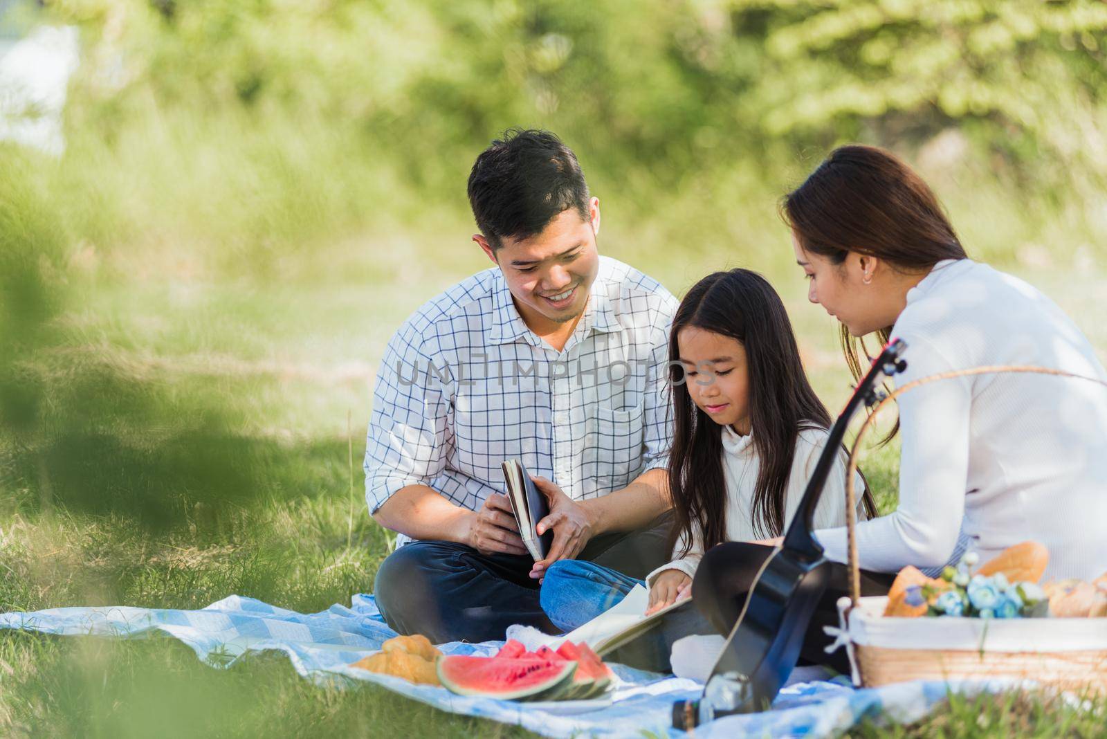 Asian family having fun and enjoying outdoor on picnic blanket reading book in park by Sorapop