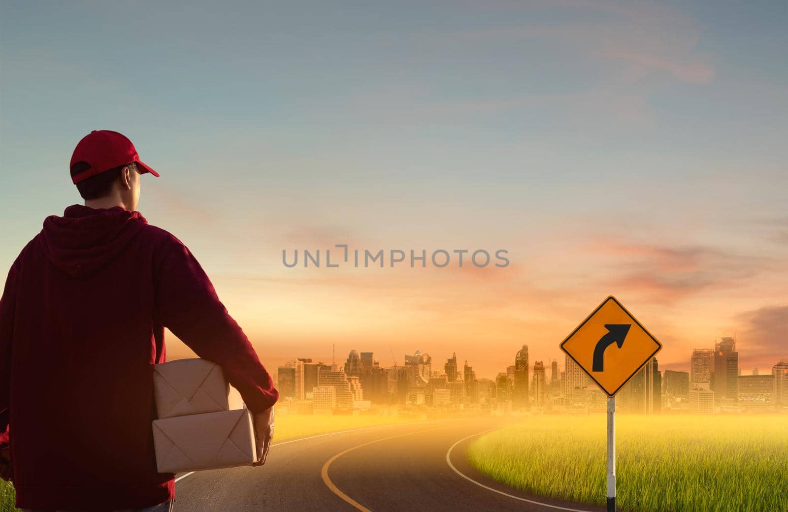 Delivery man holding cardboard boxes on the curved road at the natural sunset over field or meadow with city background.Online Shopping and Express delivery Concept. by thanumporn