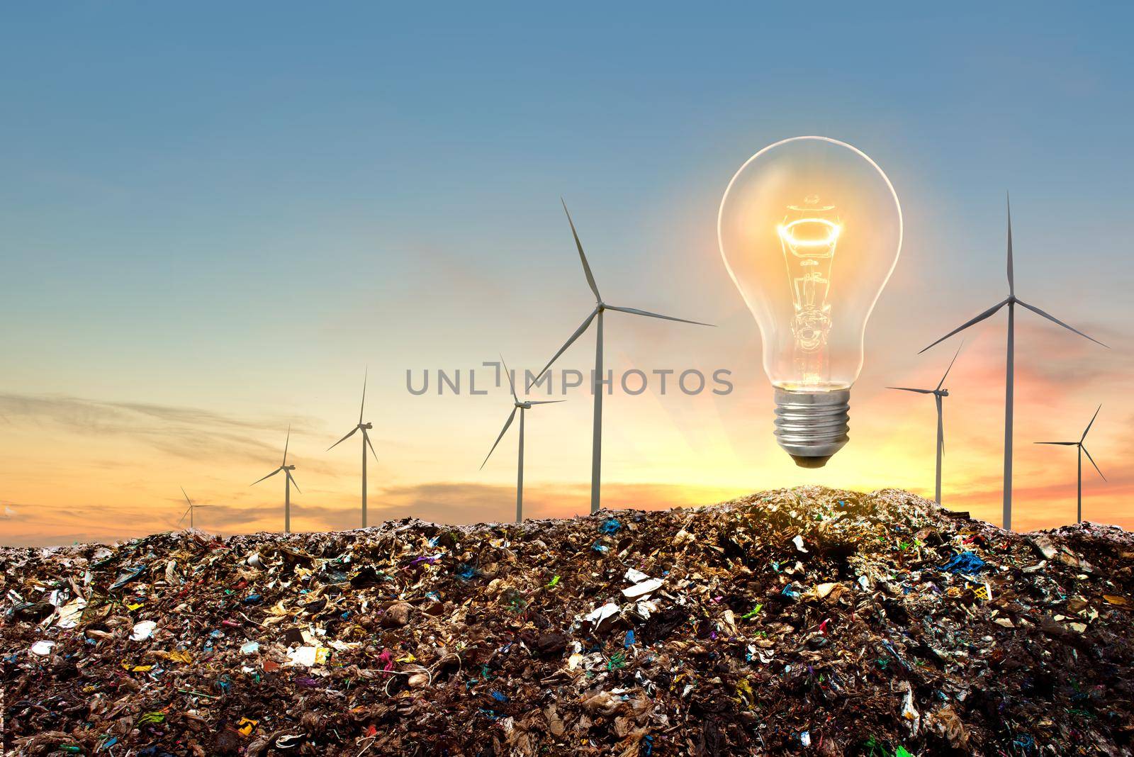 Wind turbines and lightbulb on top of garbage mountains with dramatic sunset sky background. Concept for alternative energy.