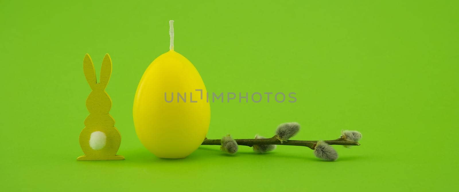 Minimalistic style Easter banner with yellow egg shaped candle, pussy willow branch and Easter Bunny figure over a green background with free copy space for your text