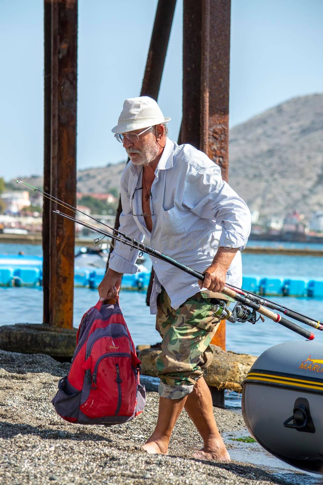 An old man with a gray beard with fishing rods near an inflatable rubber boat on the sea. Crimea, Sudak - 10 October 2020. by Essffes