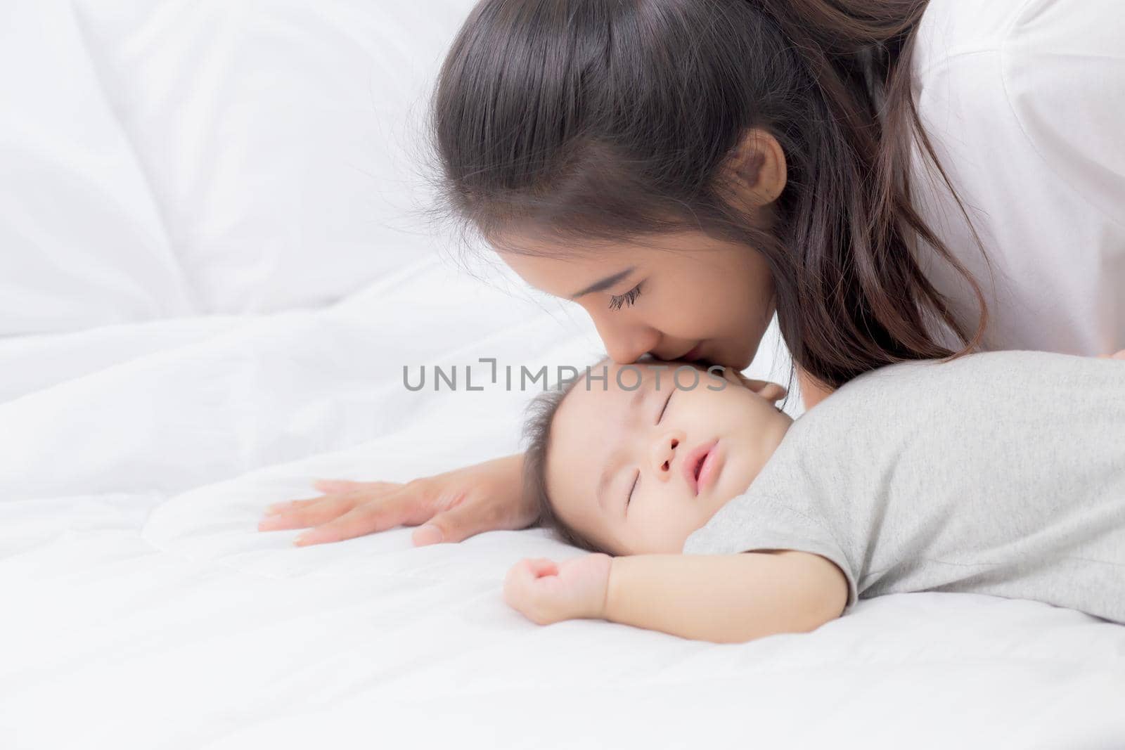 Young asian mother kiss cheek of little baby girl with tender on bed in the bedroom, mom love newborn and care, mother with expression with child together, parent and daughter, family concept.