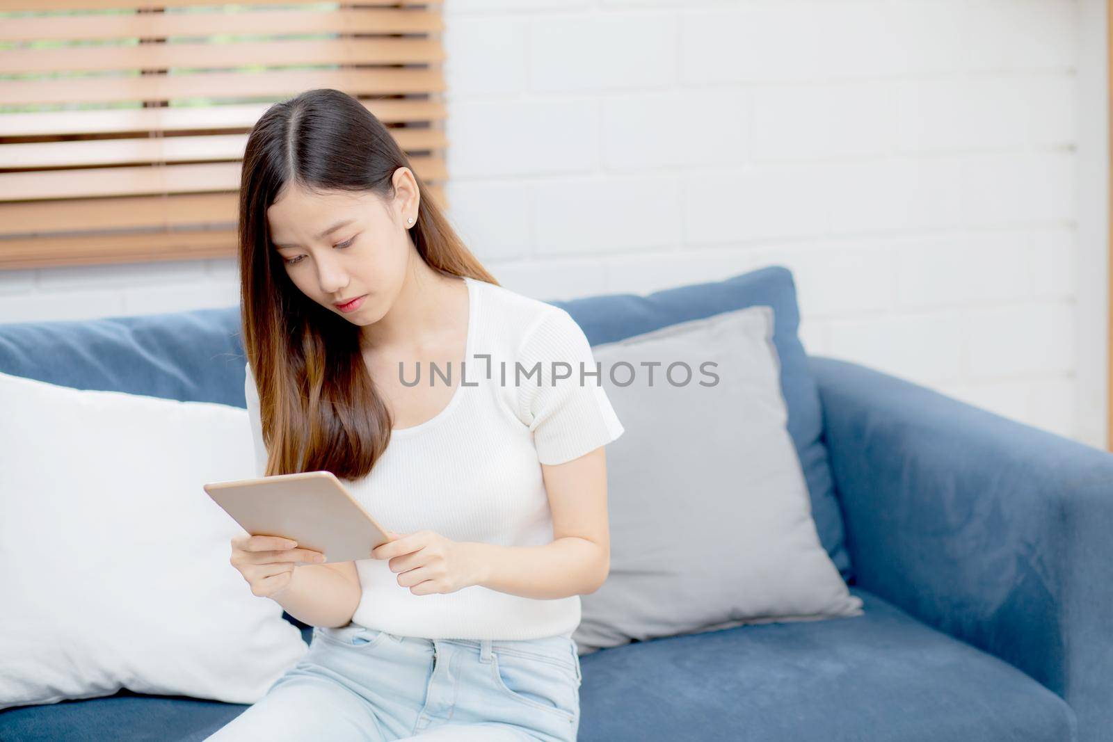 Young attractive asian woman resting using browsing tablet computer on sofa at home, happy girl sitting on couch relax reading digital gadget at house, communication and lifestyle concept.