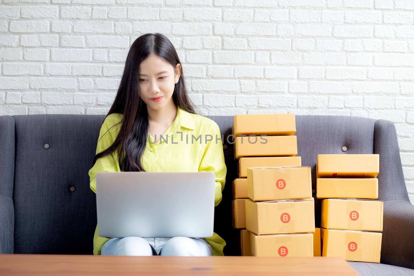 Young asian woman is merchant using laptop computer on sofa checking order of customer with purchase online shopping, business SME or startup, entrepreneur or seller and service delivery product.