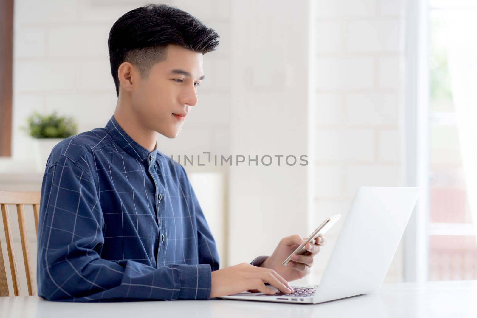 Young asian man working laptop computer and reading smartphone on internet online on desk at home, freelance male smiling using phone with social media on table, business and communication concept.