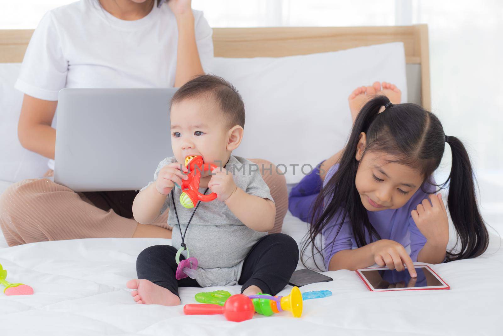 Young asian mother working at home online to internet with laptop computer and talking phone and daughter looking tablet on bed at bedroom, business woman is freelance, new normal, family concept.