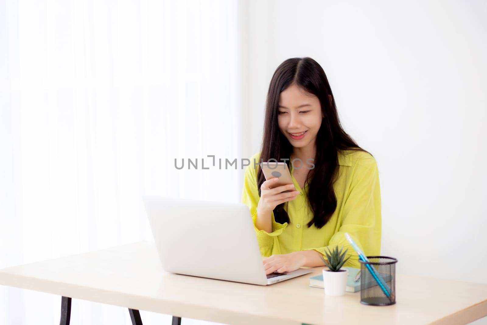 Young asian businesswoman working on laptop computer online on desk at home office, freelance woman looking message on smart phone and typing on notebook on table, business and communication concept.