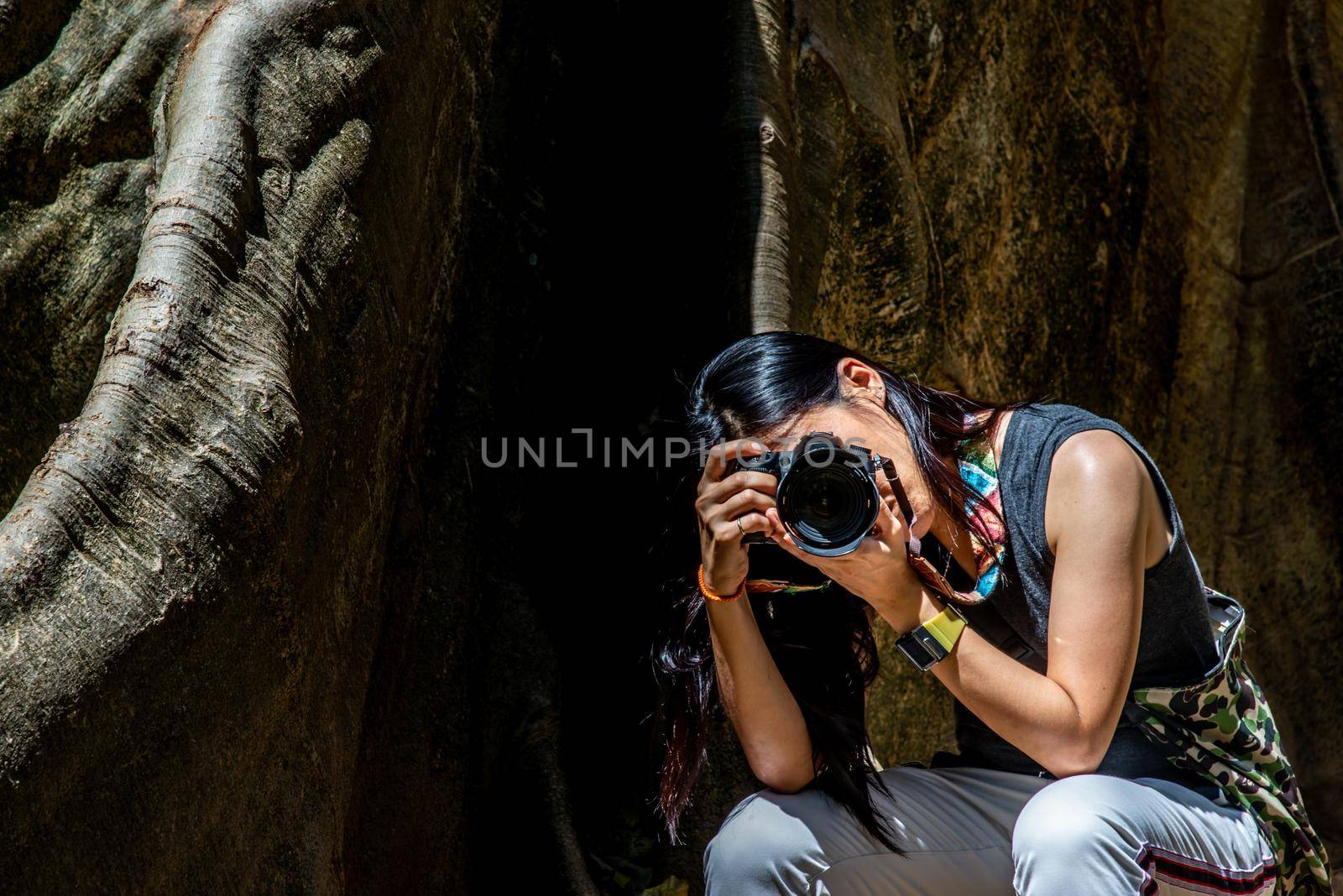 Young woman with Shoulder bag and using a camera to take photo Giant big tree, Size comparison between human and giant big tree in Ban Sanam of Uthai Thani Province, Thailand, nature background.