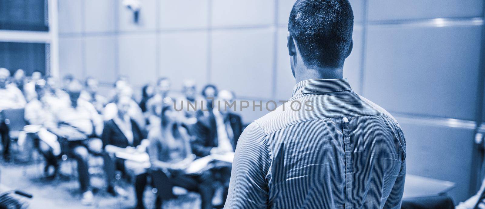 Speaker giving a talk on corporate business conference. Unrecognizable people in audience at conference hall. Business and Entrepreneurship event. Blue toned black and white.