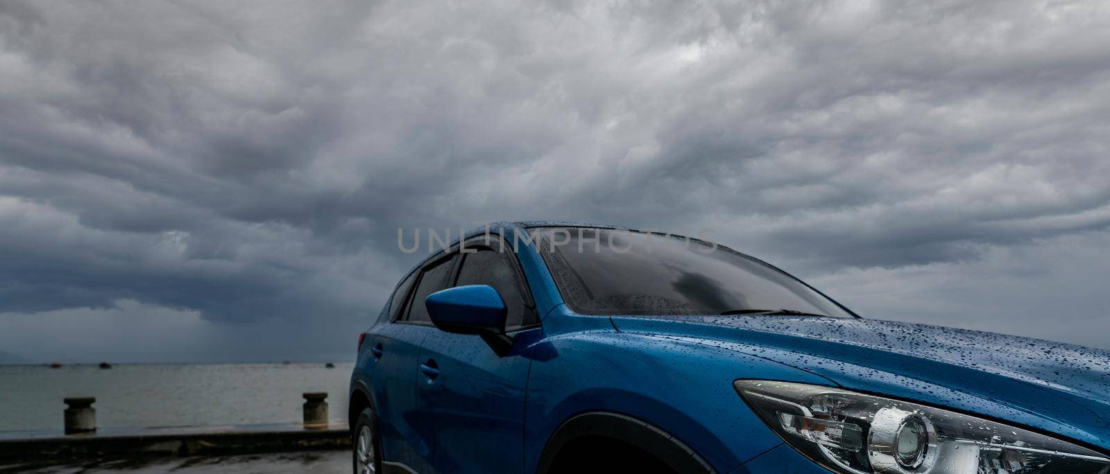 Blue SUV car with water drops parked at parking lot near sea beach against stormy and cloudy sky. Front view of new luxury SUV car with sport design. Rent car for road trip. Car driving on rainy day. 