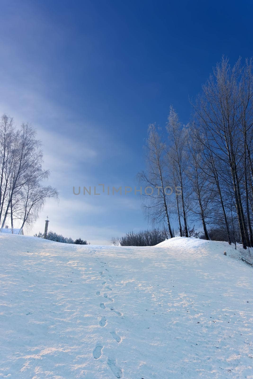 Row of footprints down a mountain slope covered in fresh white snow on a beautiful cold sunny winter day with blue sky looking up from below to trees and the roof of a house on the horizon
