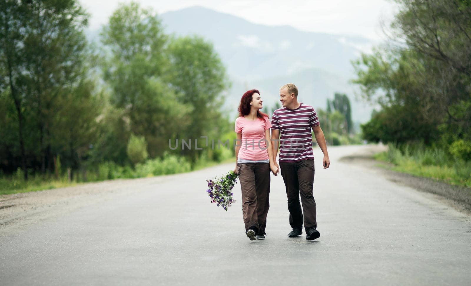 Young happy couple man and woman are walking along an empty road, the girl is holding a bouquet of flowers. 