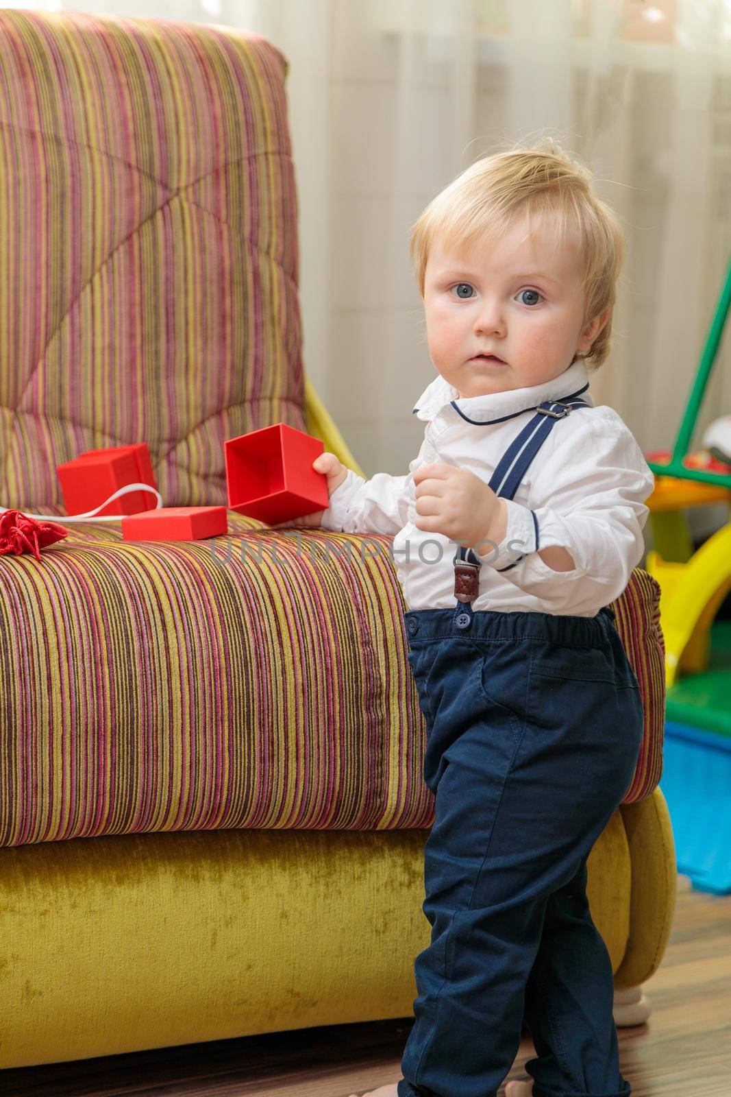 Little boy, child stands by the couch with a festive gift box. In a nice outfit, a white shirt and trousers with suspenders.