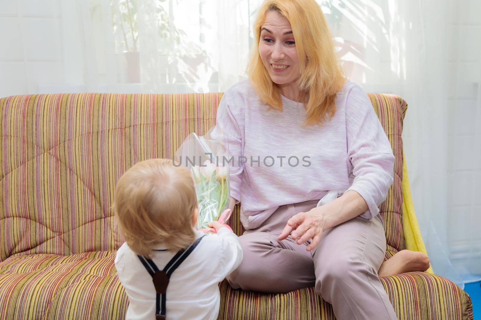 The kid congratulates his mother on the holiday, gives her flowers and a gift. Boy in white shirt and trousers with suspenders