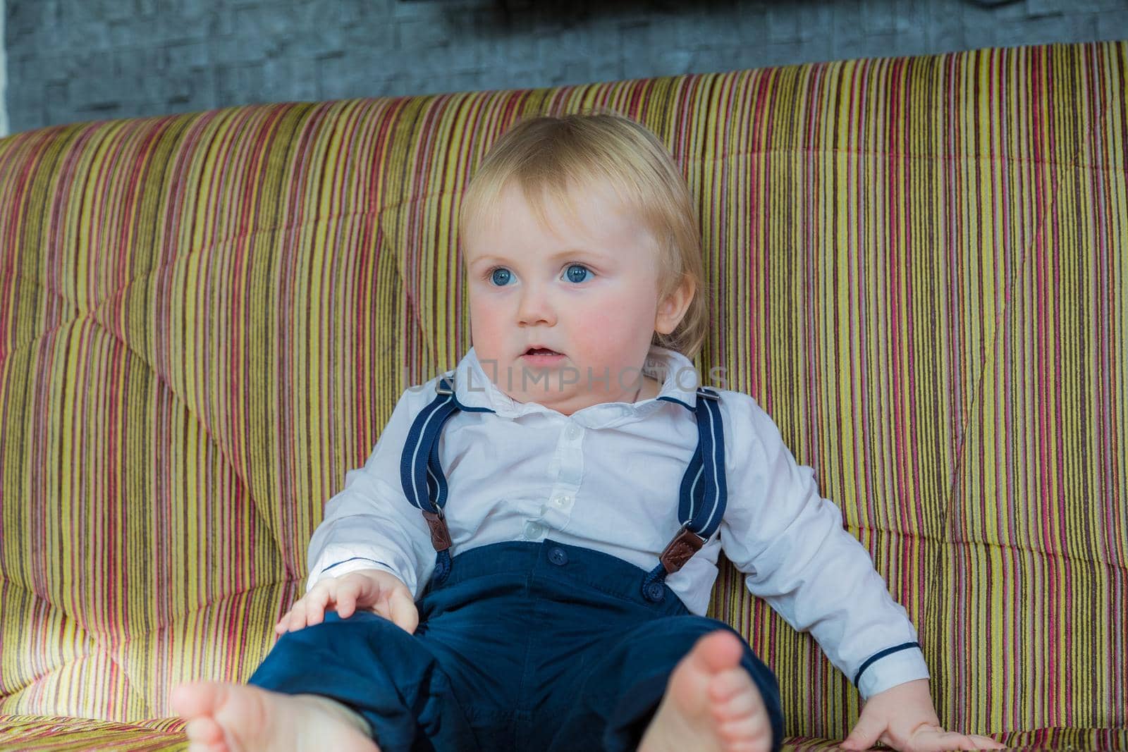 A little boy, a child, sits on the couch and waits for his parents. In a nice outfit, a white shirt and trousers with suspenders.