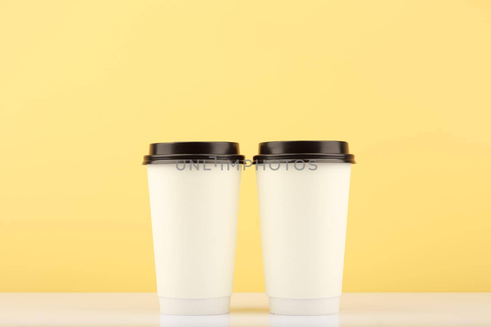 Two white tall disposable coffee or tea cups on white table against light yellow background with copy space. Hot drinks minimalistic concept