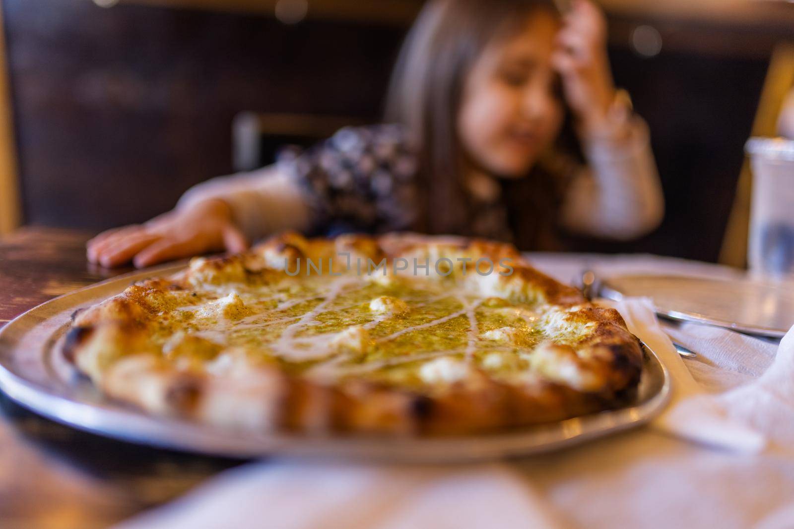 Tasty cheese and species pizza on vintage-looking wooden table with blurry little girl as background. Young child at rustic table ready to eat delicious Italian dish. Food and drinks