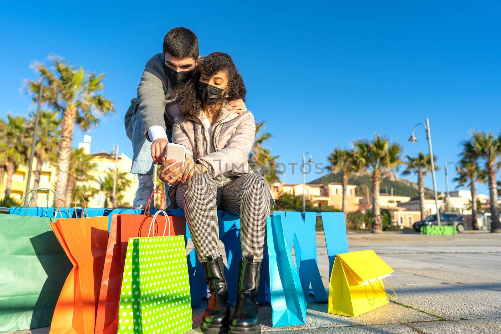 Multiracial heterosexual couple of young people outdoors sitting on a bench among the colorful shopping bags using and pointing the phone to buy online wearing the protective mask against Coronavirus.