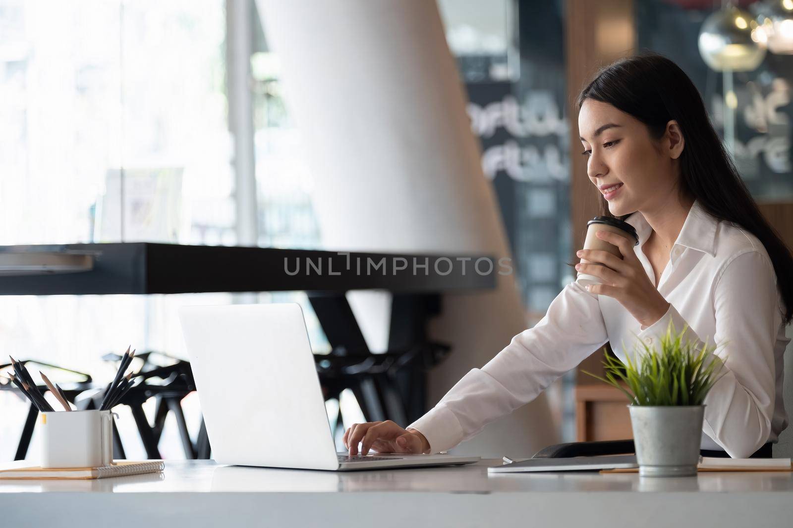 Remote work. Asian woman working remotely on her laptop. A brunette girl in a white shirt doing notes during an online business briefing at her home workplace by nateemee