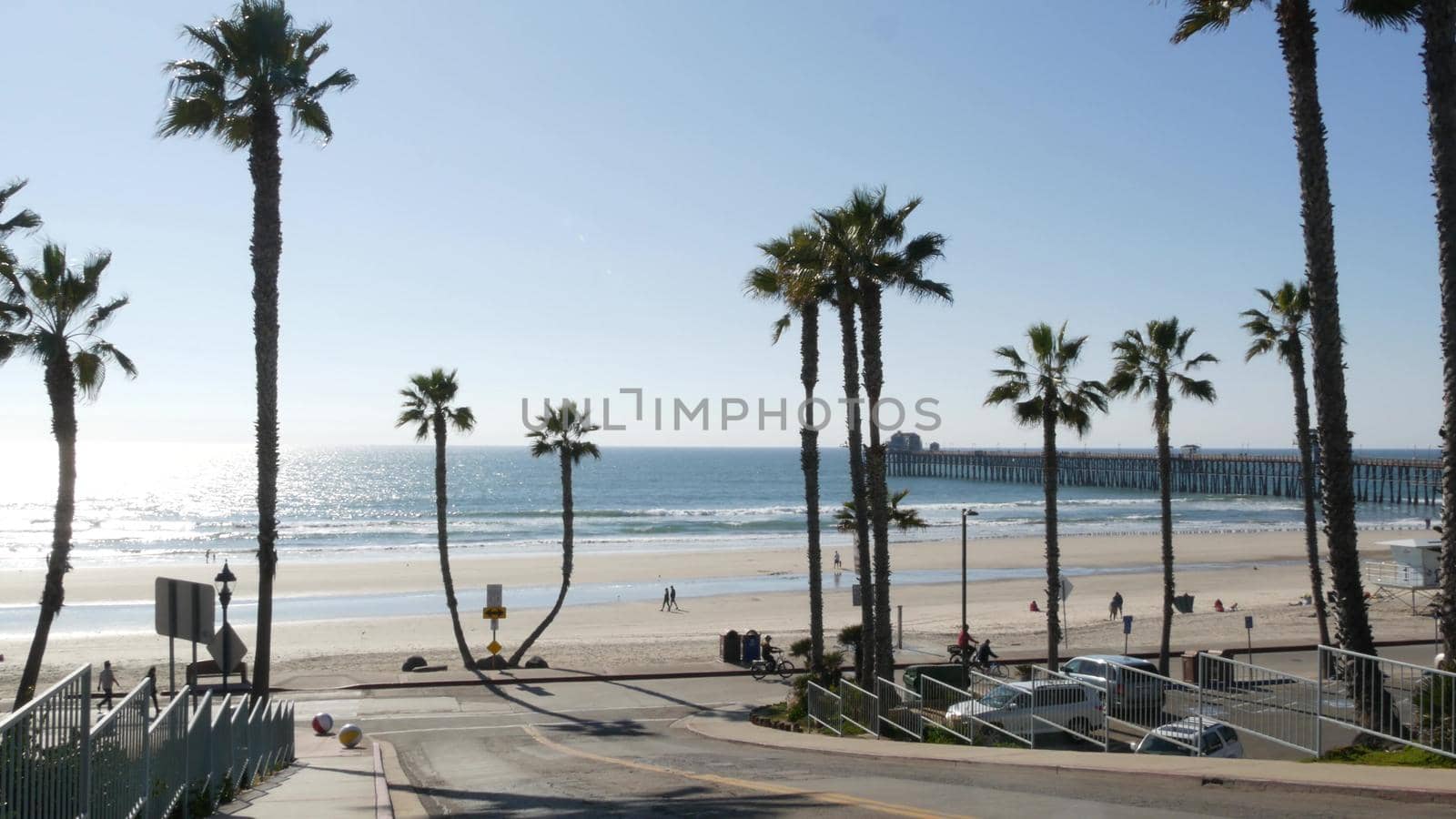 Oceanside, California USA - 8 Feb 2020: People walking strolling, waterfront resort street, pacific ocean tropical beach with palm trees. Person riding bicycle, coastal road perspective. Biker cycling